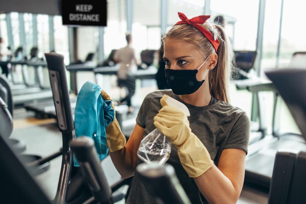 A young woman wearing a mask and a pair of cleaning gloves cleaning an elliptical trainer by spraying with a cleaning solution and wiping it down with a blue microfibre cloth