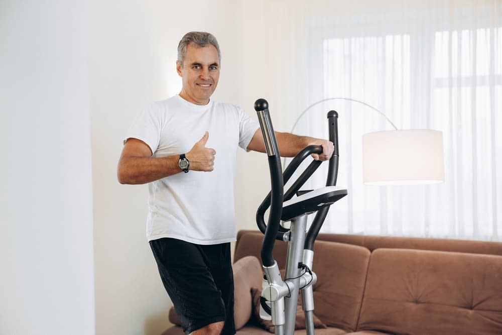 Middle-aged man posing thumbs up on his elliptical machine in his living room