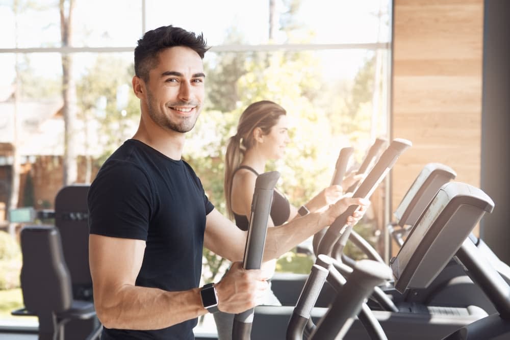 A friendly looking young man exercising on an elliptical machine, with a female companion doing the same thing in the background